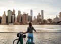 Individual standing by a railing with a bicycle, gazing at the New York City skyline across the water.